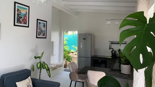 Een moderne woonkamer met een bank, stoelen, een eettafel en een koelkast. Twee ingelijste foto's hangen aan de muur, en er staat een potplant en een grote, weelderige plant op de voorgrond.