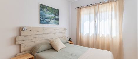 Un dormitorio con una cama, una mesita de noche, un aire acondicionado de pared, un cuadro y una ventana con cortinas.