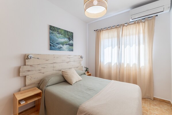 A bedroom with a bed, a nightstand, a wall-mounted air conditioner, a painting, and a window with curtains.
