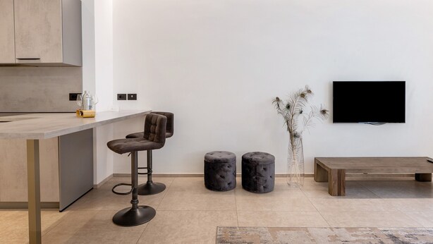 A modern kitchen with a bar stool, two ottomans, a bench, and a flat-screen TV mounted on the wall.