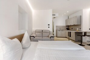 A modern hotel room with a bed, a sofa, a kitchen area, and a bar.