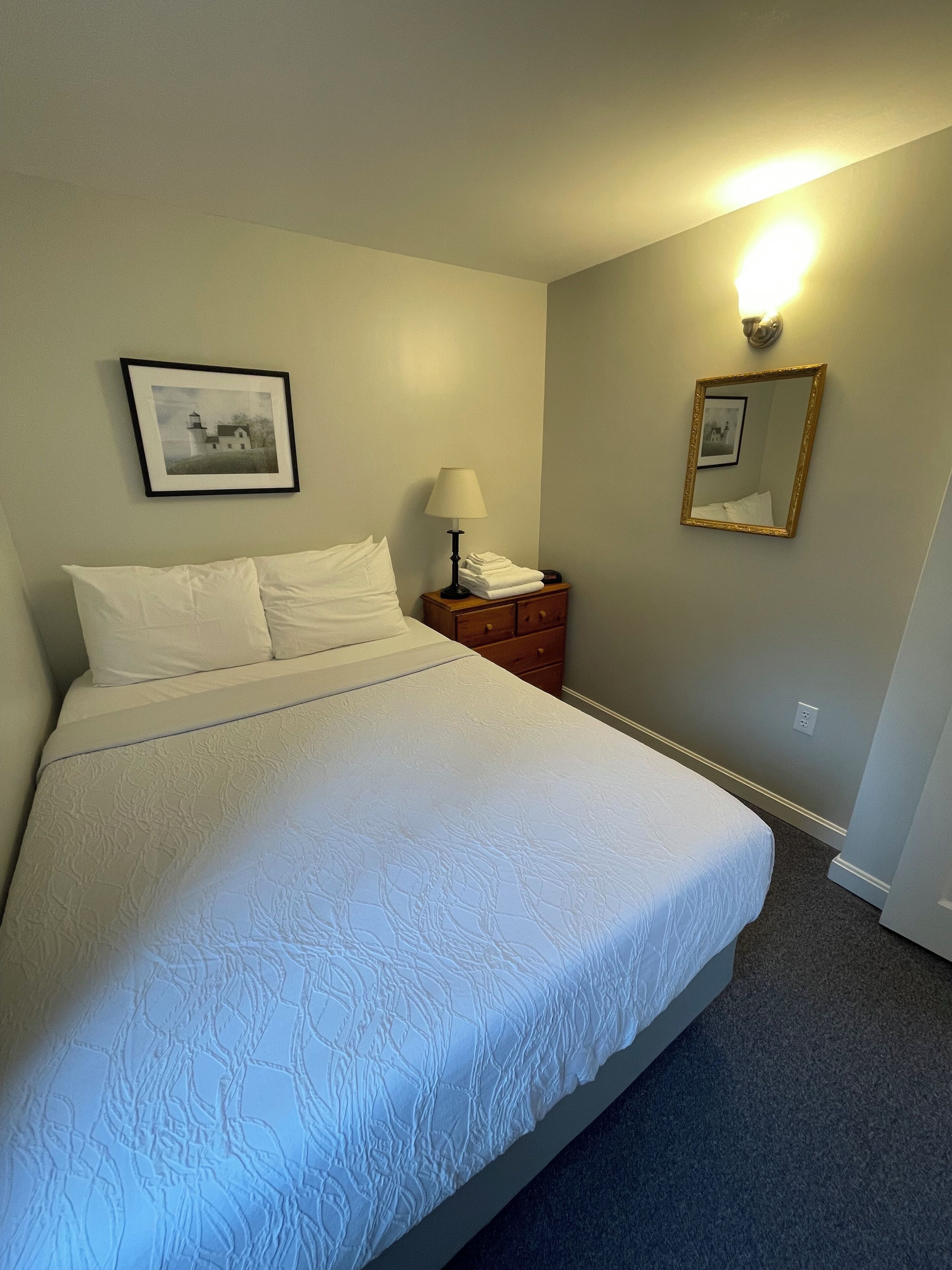 A neatly made bed with a white bedspread, a bedside lamp, a framed picture on the wall, and a mirror.