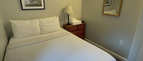 A neatly made bed with a white bedspread, a bedside lamp, a framed picture on the wall, and a mirror.
