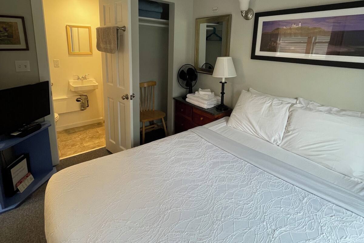 A hotel room with a bed, a desk with a lamp, a chair, a wardrobe, and a television.