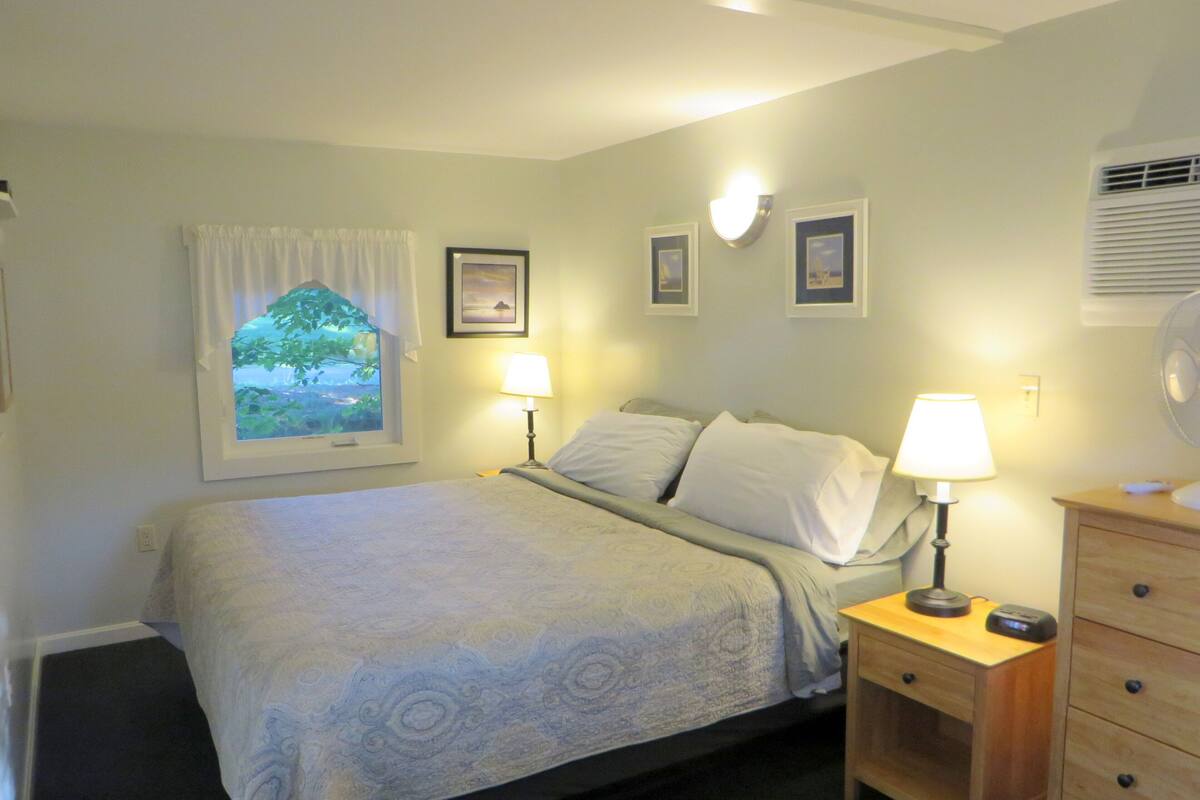 A bedroom with a bed, two bedside tables, a dresser, a window with curtains, and wall-mounted lamps.