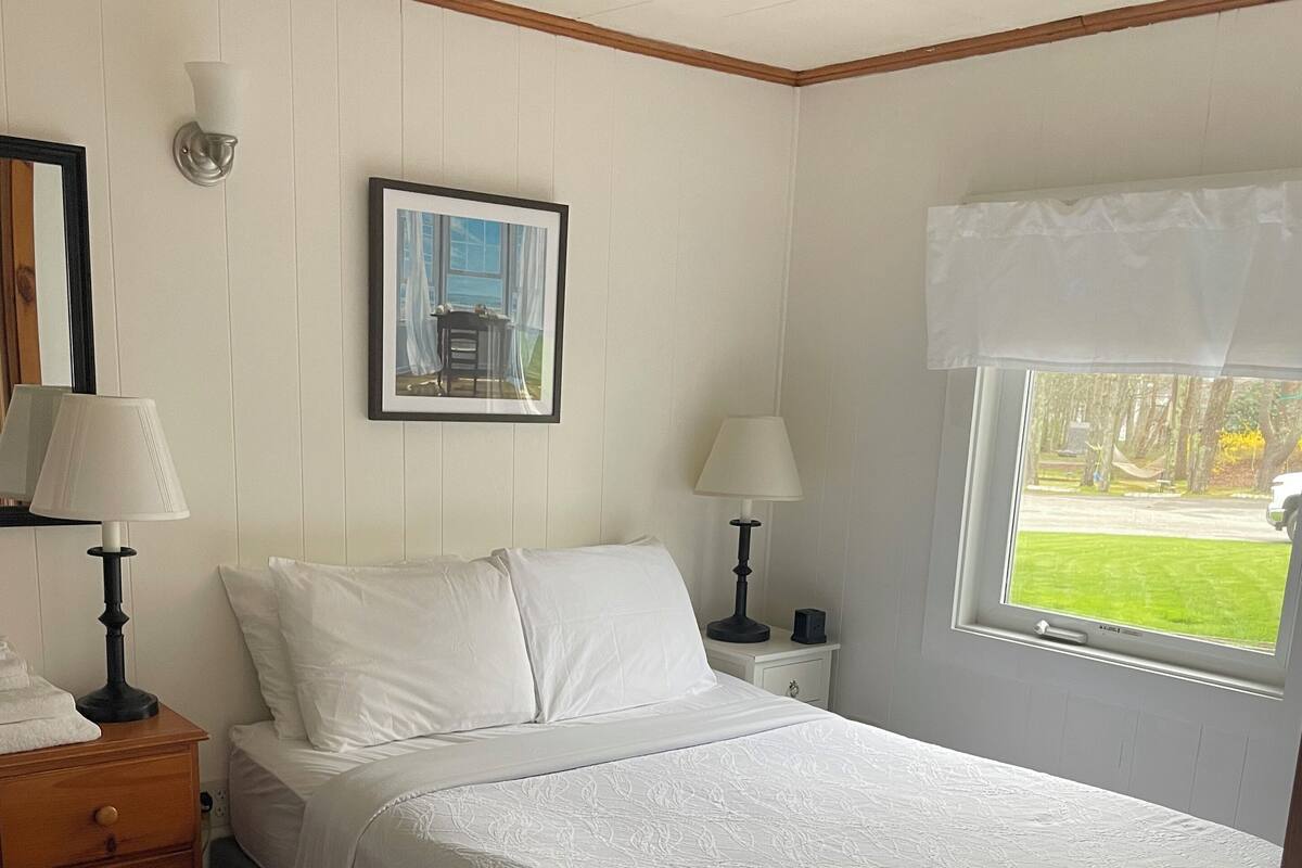 A neatly made bed in a room with a window, a framed picture on the wall, and a nightstand with a lamp.