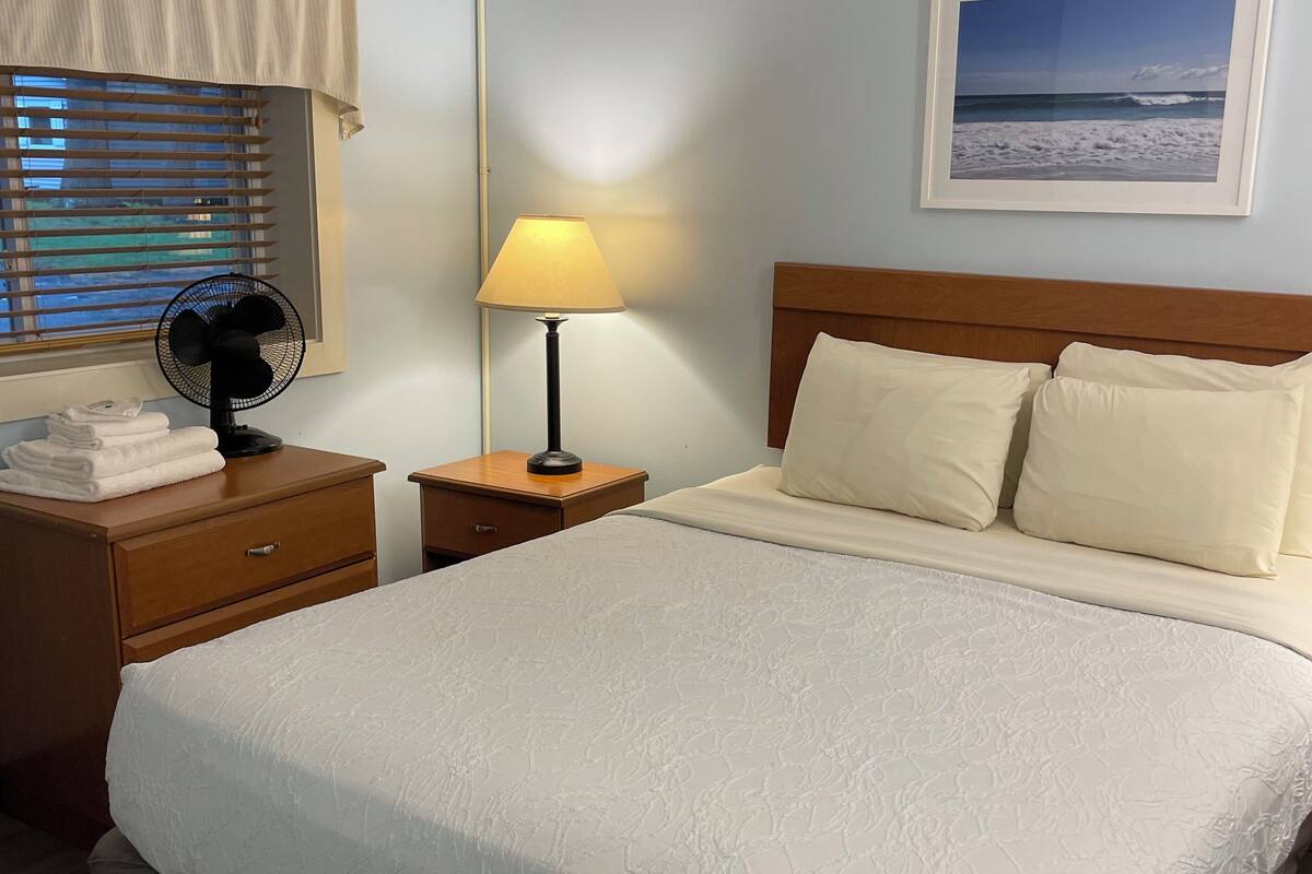 A neatly made bed with a white comforter and pillows, a bedside table with a lamp, a fan, and folded towels.
