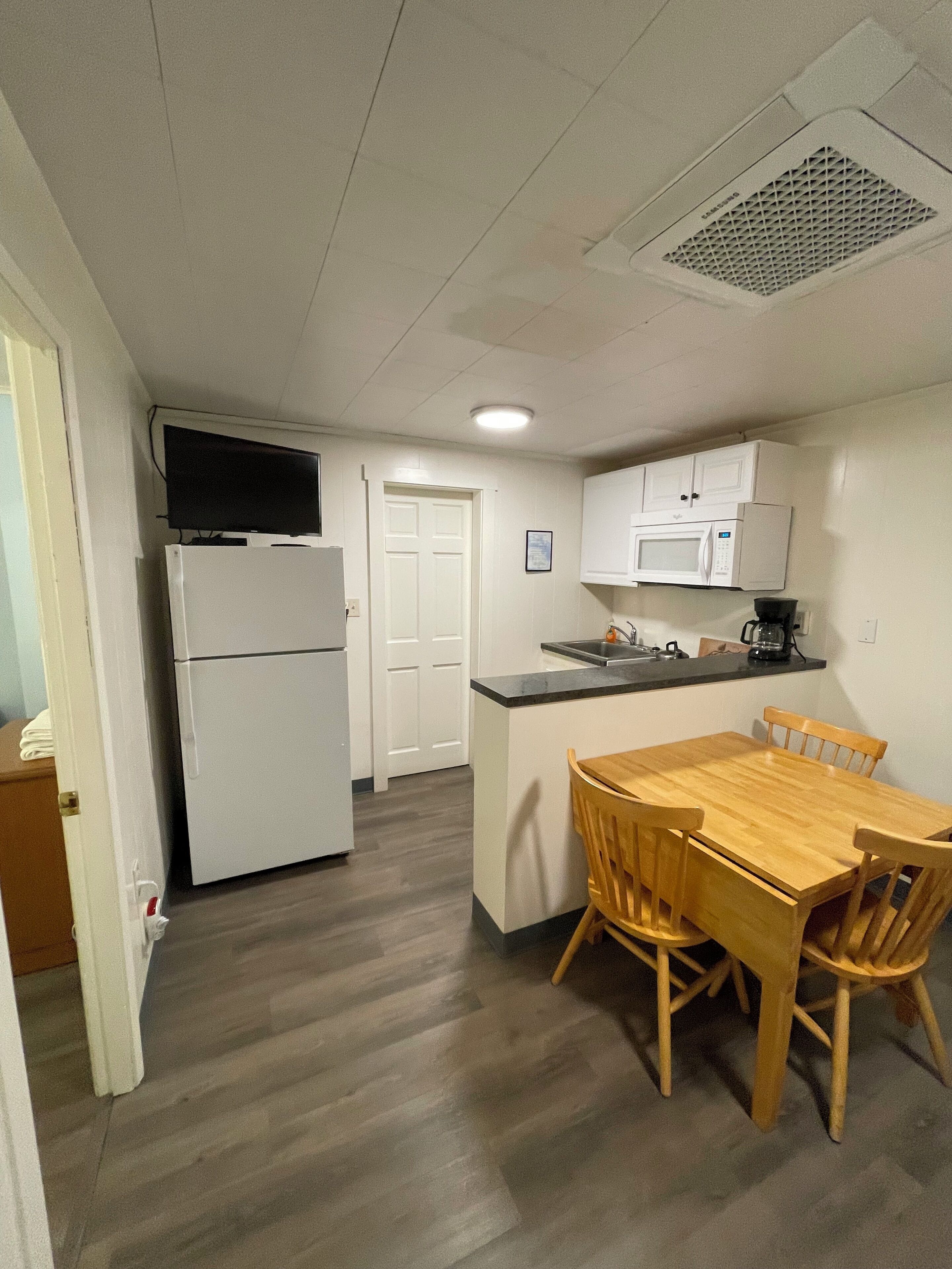 A compact kitchen with a refrigerator, microwave, and coffee maker. There is a dining table with chairs and a door leading to another room.