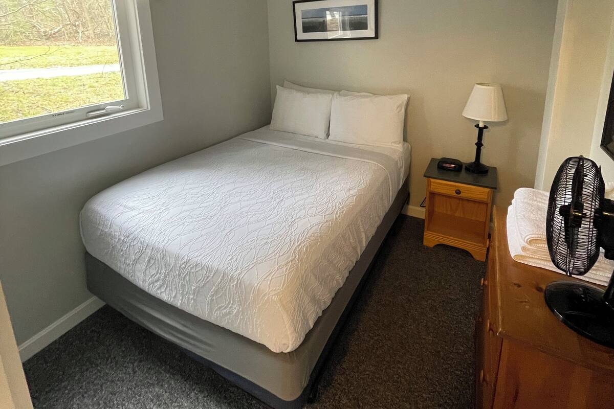 A bedroom with a single bed, a nightstand with a lamp, a fan, and a framed picture on the wall.
