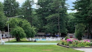 Two white Adirondack chairs facing a swimming pool with a garden of flowers and a row of beach chairs. - Elmwood Motor Court (Old Orchard Beach)
