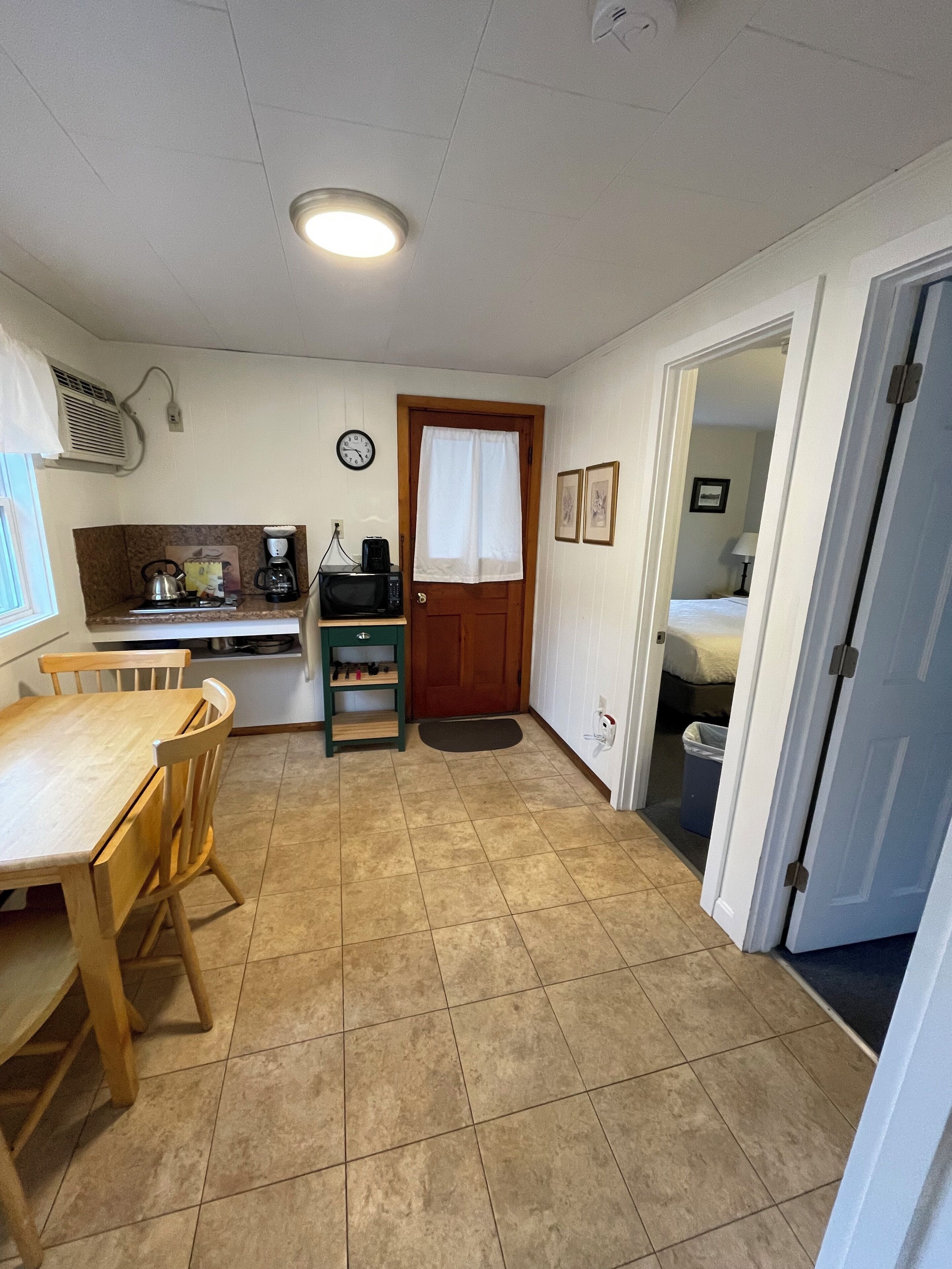 A compact kitchen with a dining table and chairs, a microwave, a kettle, and a clock on the wall.