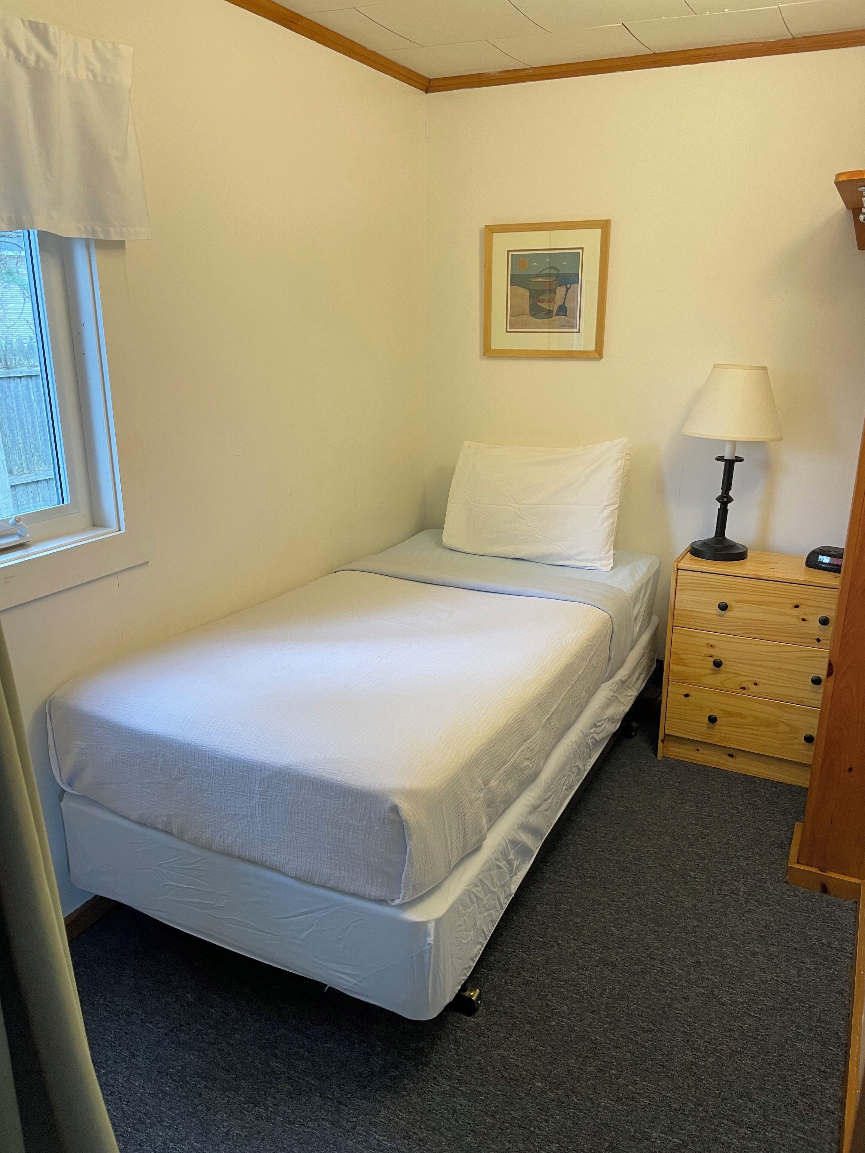 A small, neatly made bed in a room with a window, a bedside table with a lamp, and a wooden dresser.