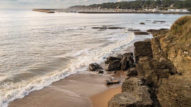 A coastal beach with rocky shoreline, calm sea, and a distant marina.