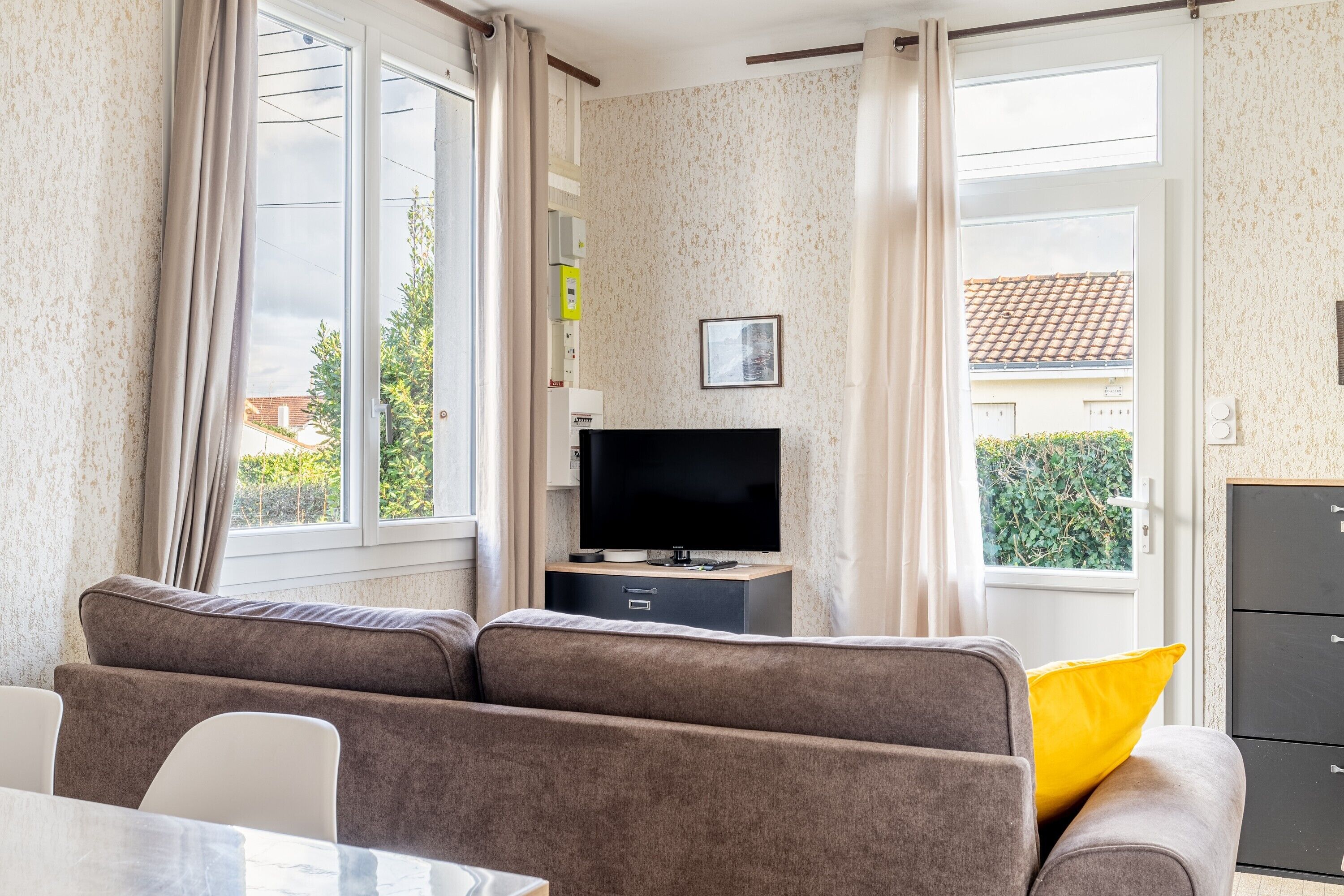 A living room with a sofa, a TV on a stand, a dining table, and a view through a window.