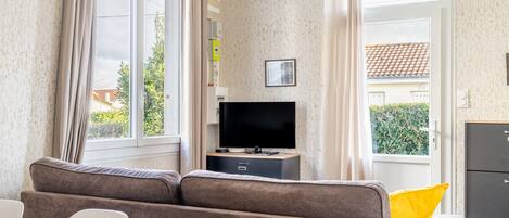 A living room with a sofa, a TV on a stand, a dining table, and a view through a window.