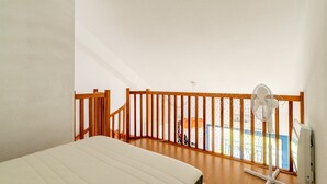 Ein Zimmer mit einem Bett, einem Standventilator, einer Holzwand und Blick auf einen Balkon.