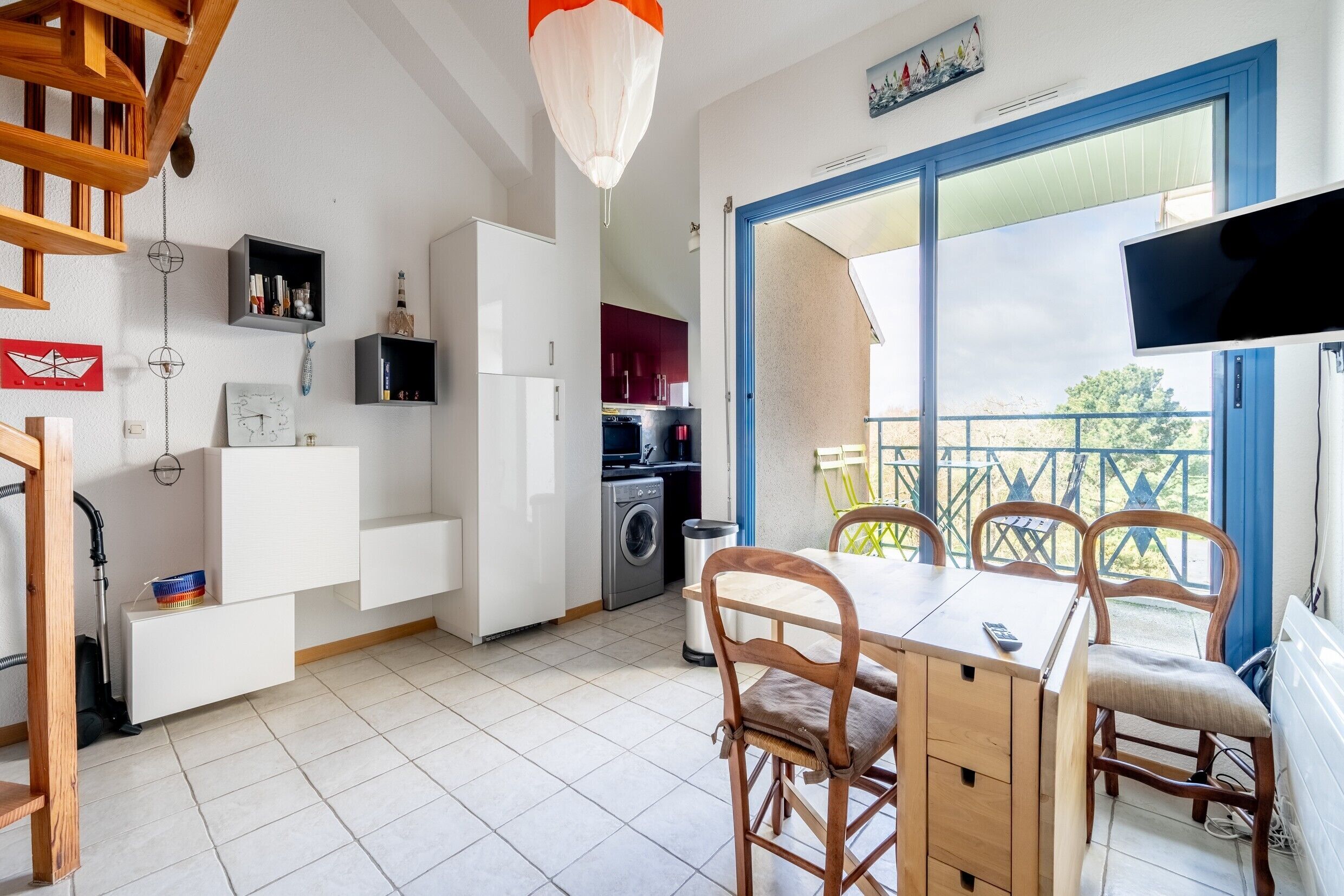 Una cocina moderna con armarios blancos, un comedor con sillas de madera, un televisor montado en la pared y un balcón con vistas.