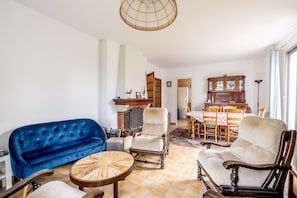 Un salon avec un canapé bleu, deux fauteuils, une table basse en bois et un coin repas comprenant une table et des chaises en bois.