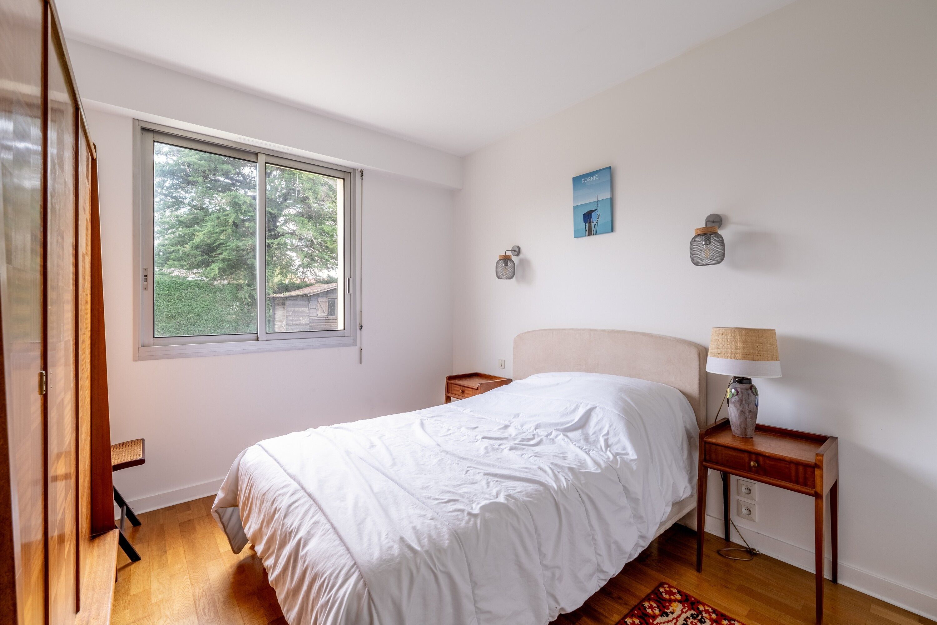 Une chambre à coucher avec un lit, une table de chevet, une lampe, une fenêtre et un tableau au mur.