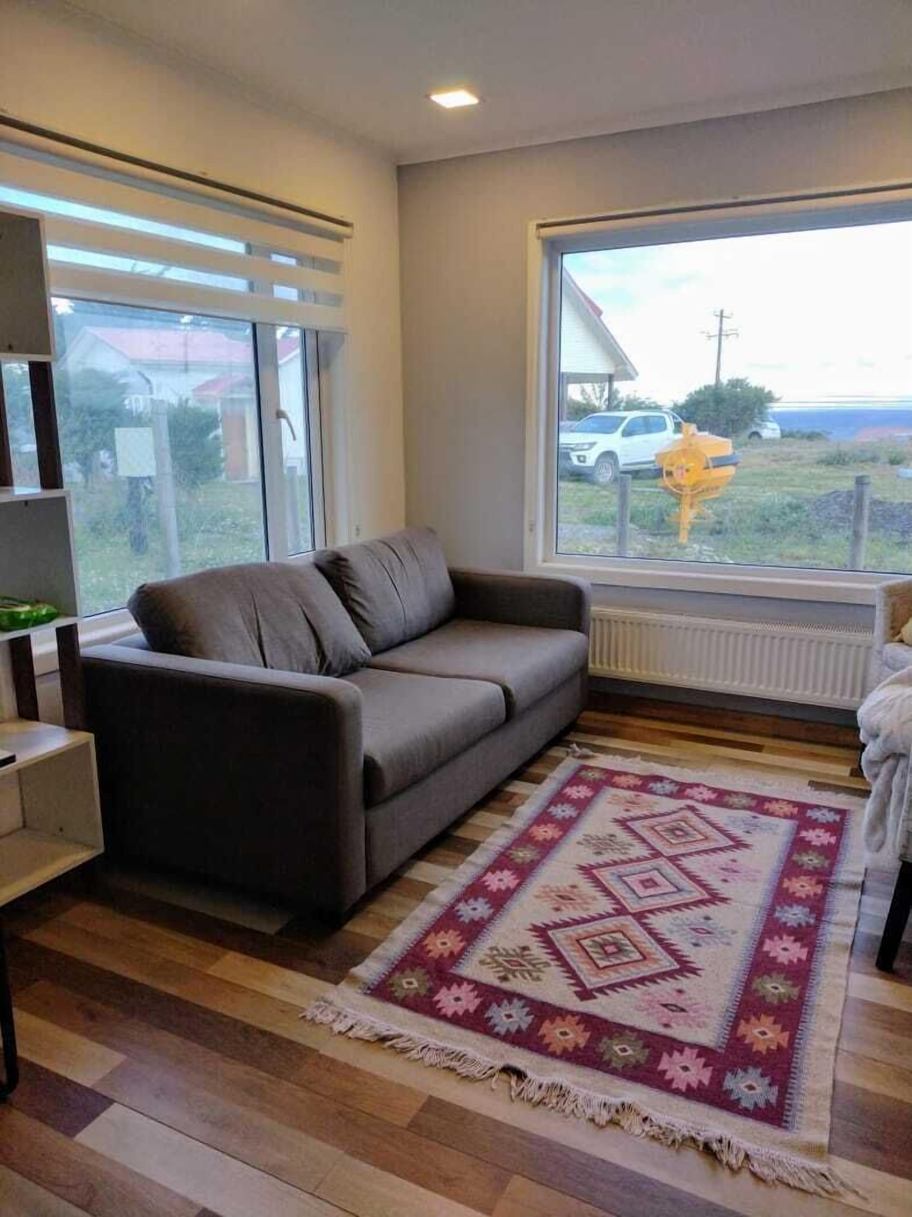 A modern living room with a grey sofa, a patterned rug, and a large window offering a view of the outdoors.