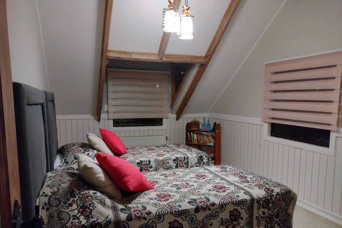 Une chambre avec un plafond en pente, un lit recouvert d’une literie à motifs floraux, une petite bibliothèque et une fenêtre avec des stores.