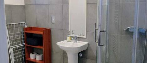 Een badkamer met een wit zuilwastafelmeubel, een spiegel, een douchecabine, een houten plankje met toiletartikelen, en vloertegels met een patroon.