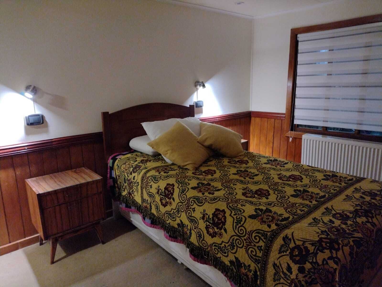 A bedroom with a wooden bed, two bedside tables, a window with blinds, and a wall-mounted light fixture.