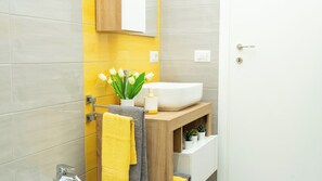 Un baño con un lavamanos blanco, toallas amarillas y grises, un tocador de madera y un florero con flores blancas.