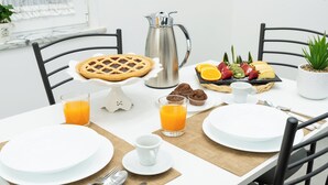 Una mesa de desayuno dispuesta con platos, vasos, una cafetera, muffins, pastel y una bandeja de frutas.