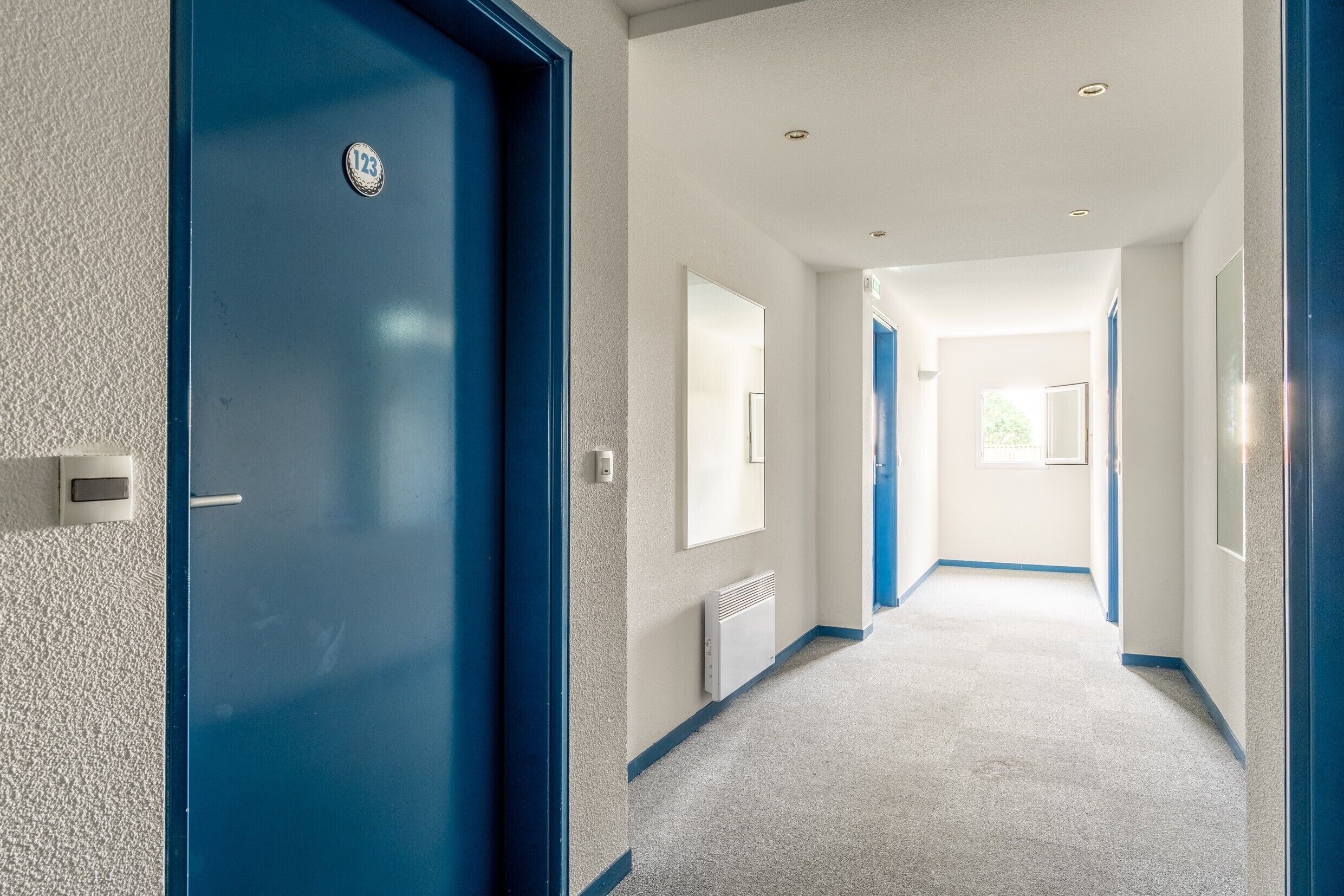 Un couloir avec une porte bleue, un interrupteur, une grille d’aération et une fenêtre au fond.