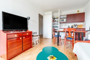A compact hotel room with a TV, a wooden dresser, a dining table with chairs, a kitchen area, and a bed.