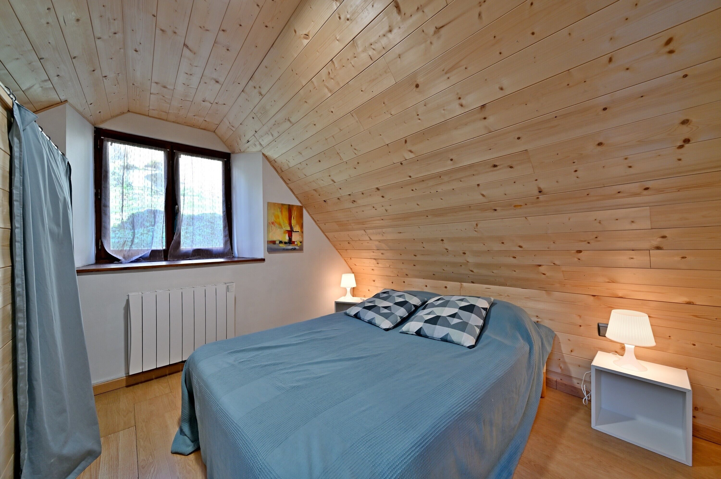 A bedroom with a sloped ceiling, a bed with a blue bedspread, two bedside lamps, a window with curtains, and a radiator.