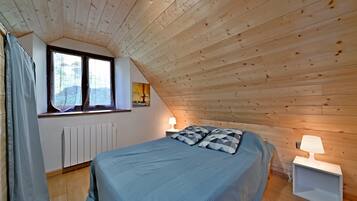 A bedroom with a sloped ceiling, a bed with a blue bedspread, two bedside lamps, a window with curtains, and a radiator.