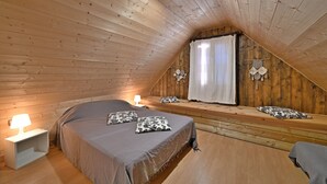Un dormitorio acogedor en un ático con techo inclinado, paredes de madera, cama con almohadas, lámpara de noche y ventana con cortinas.
