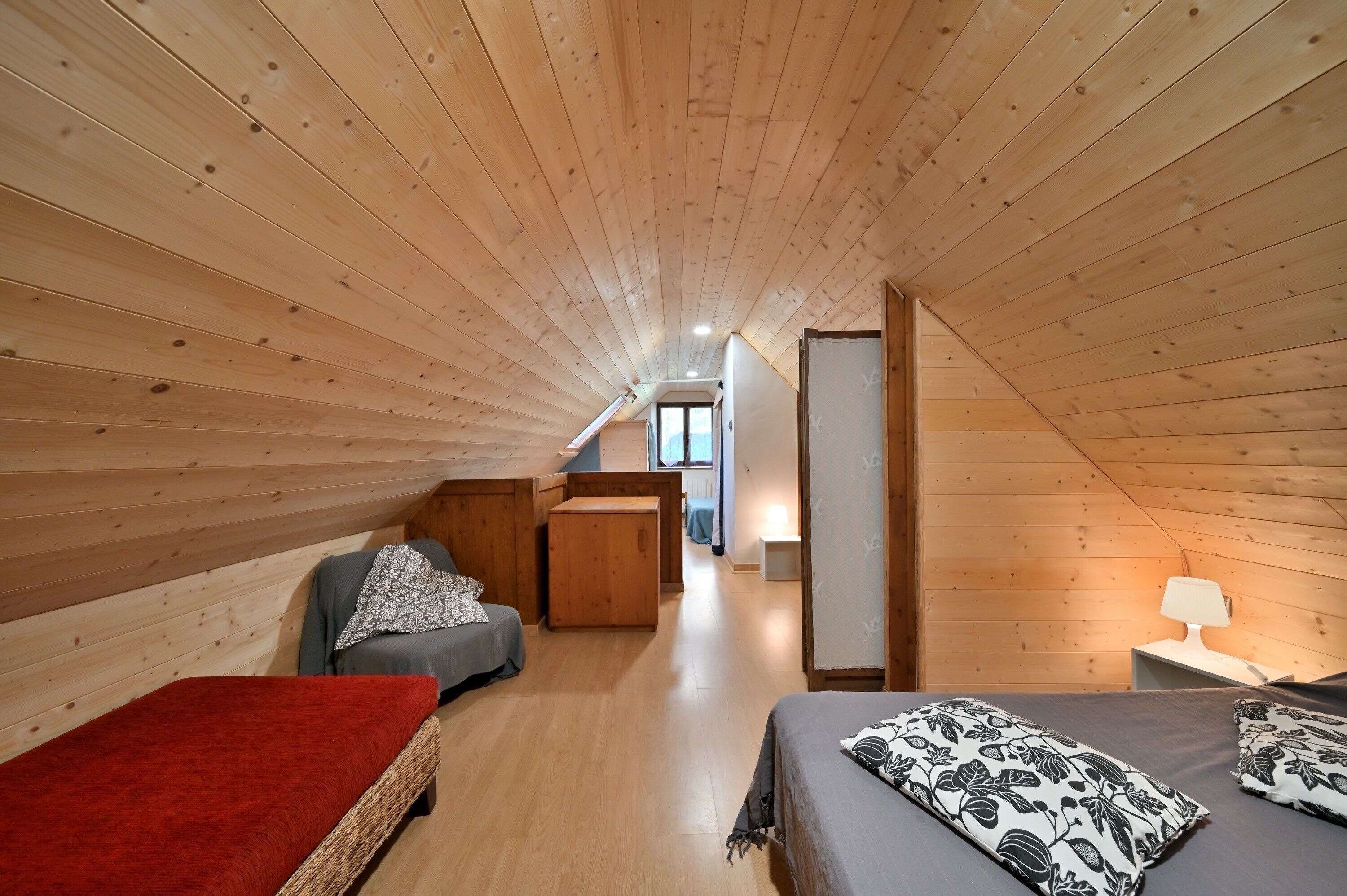 Une chambre mansardée aux murs lambrissés, avec un lit, un petit canapé, un banc rouge et une table de chevet avec une lampe.