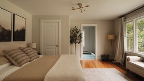 Un dormitorio con una cama grande, una mesita de noche, una lámpara, una ventana con cortinas y una puerta que da a otra habitación.