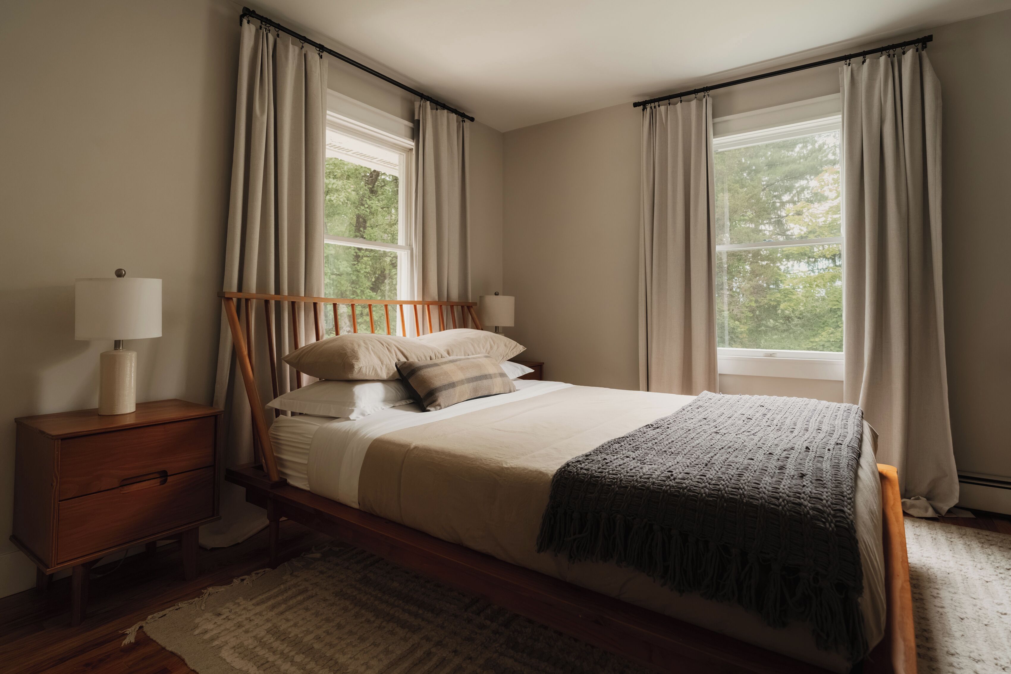 Un dormitorio con una cama de madera, una mesita de noche, una lámpara y dos ventanas con cortinas.