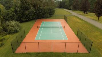 A tennis court surrounded by a fence, with a road and trees in the background.