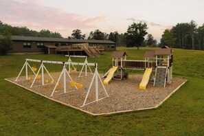 Un parque infantil con columpios y toboganes, rodeado por una valla de madera y un edificio al fondo.