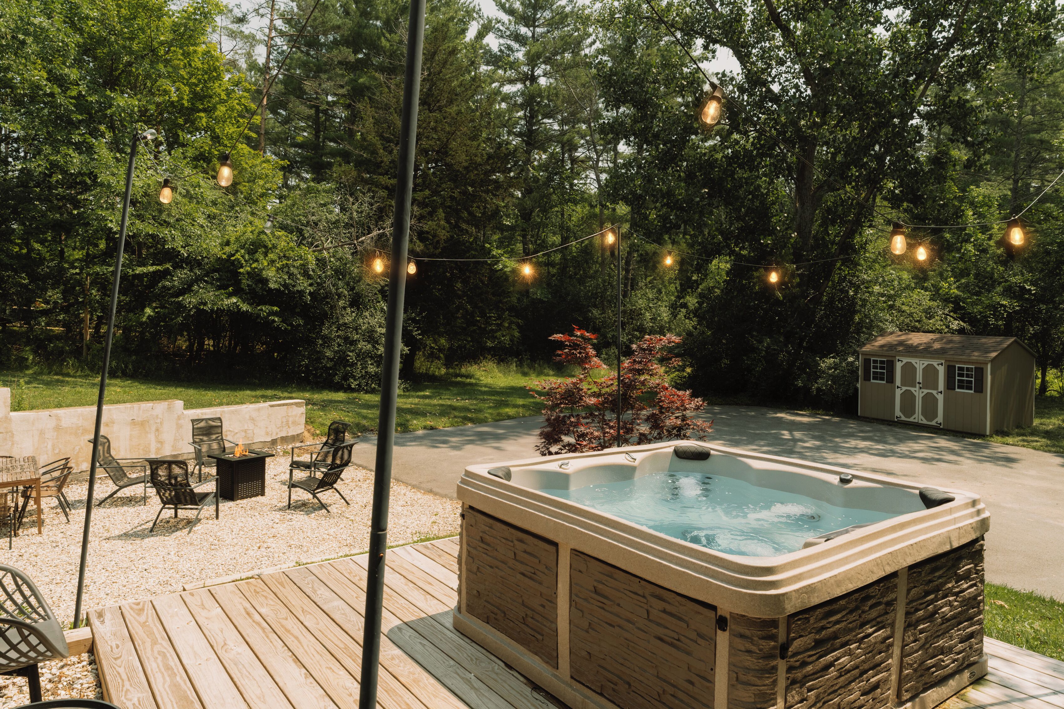 Un jacuzzi en una terraza de madera con mobiliario exterior, rodeado de árboles y un cobertizo.
