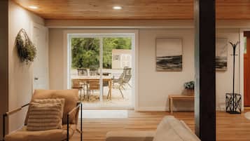 Un'ampia zona giorno con soffitto in legno, una grande porta scorrevole in vetro che dà su un patio esterno e vista su un giardino e un deposito.