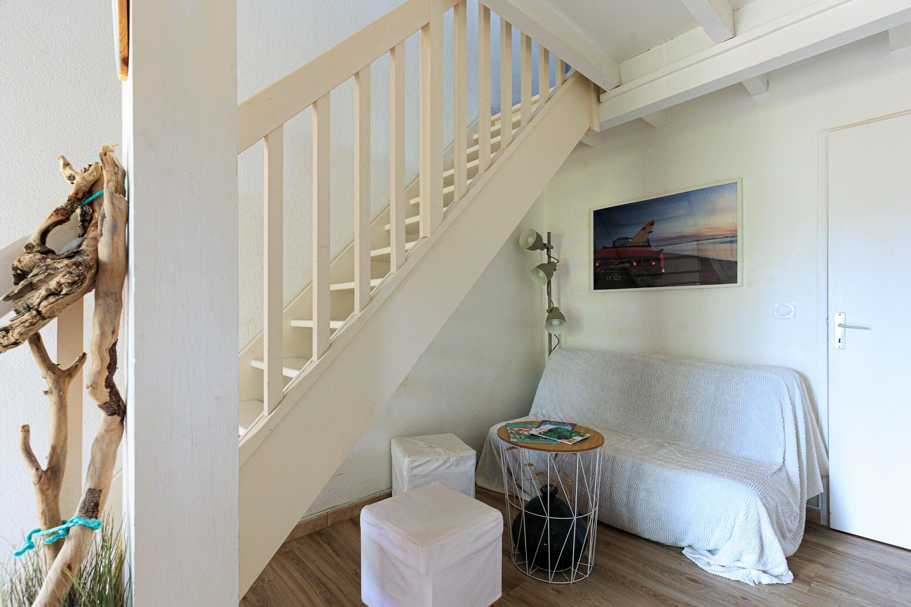 A staircase with a white railing, a bed with a white cover, a small round table with a magazine, and a door with a light switch.