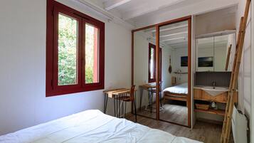 A bedroom with a bed, a desk, a chair, a mirror, and a bathroom with a sink and mirror.