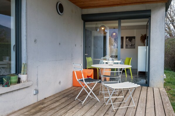 Un patio moderno al aire libre con piso de madera, puerta de vidrio que da a un espacio interior y una mesa con sillas.
