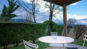 Een overdekt terras met een ronde tafel en stoelen, een glas water, en uitzicht op bomen en heuvels.
