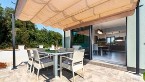An outdoor patio with a table and chairs, a large umbrella, and a view into a modern kitchen and dining area. - Amazing home in Rovinj (Rovinj)