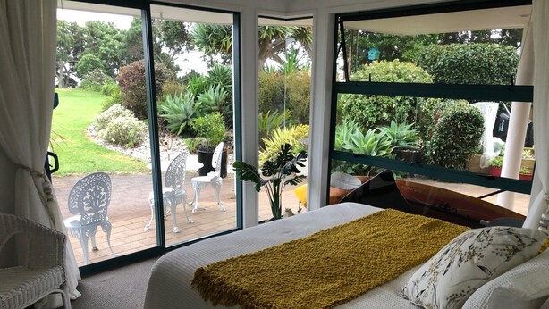 Una camera da letto con un letto grande, una coperta gialla e vista su un giardino attraverso finestre a tutta altezza.
