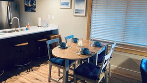 Une cuisine moderne dotée d’un évier encastré, d’un coin repas avec une table en bois et des chaises bleues, et d’une grande fenêtre avec des stores.