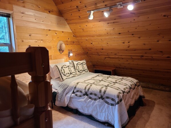 A cozy bedroom with a wooden sloped ceiling, a bed with patterned bedding, a small bedside table, and a wall-mounted light fixture.