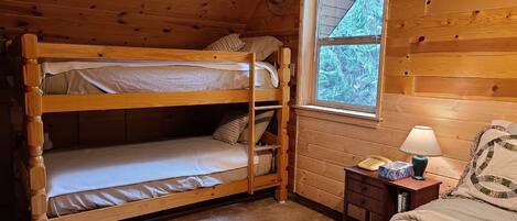 A cabin room with a bunk bed, a small bedside table with a lamp, and a window with a view of trees.